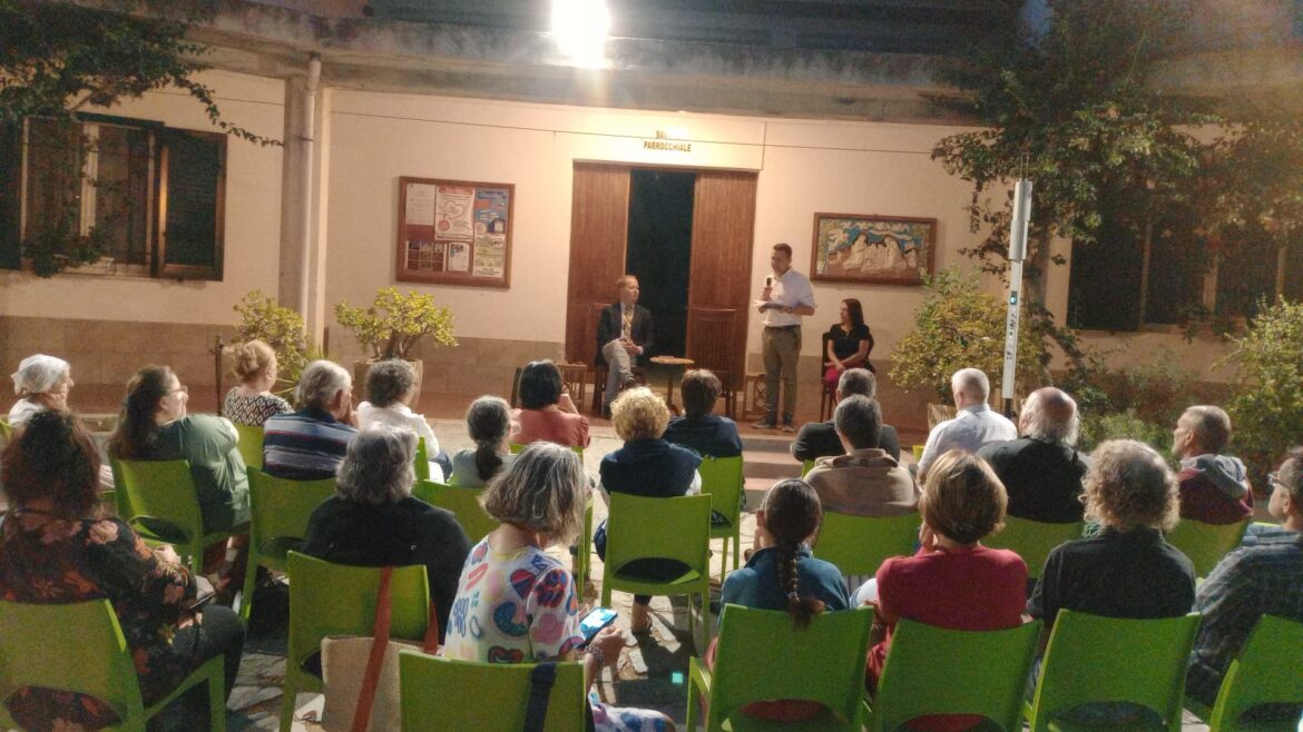 Foto della presentazione TLE a Porto Ferraio; visibili varie persone sedute, da dietro, e Mario Cignoni di fronte a loro intento a presentare la nuova traduzione.
